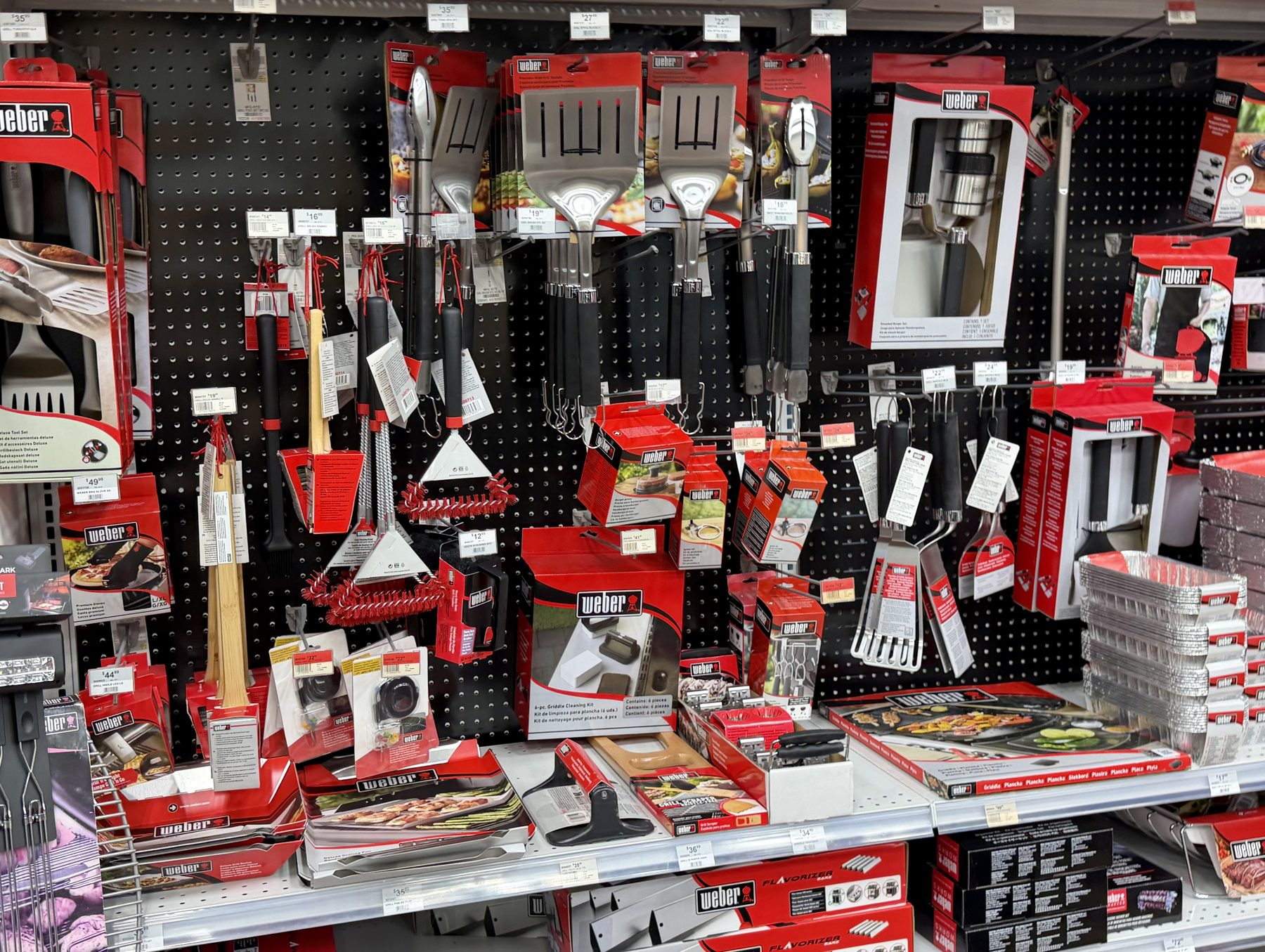 Weber grilling tools and accessories including spatulas, tongs, grill cleaning brushes, and aluminum drip pans on pegboard displays at Hessler Paint & Hardware in Delray Beach, Florida.