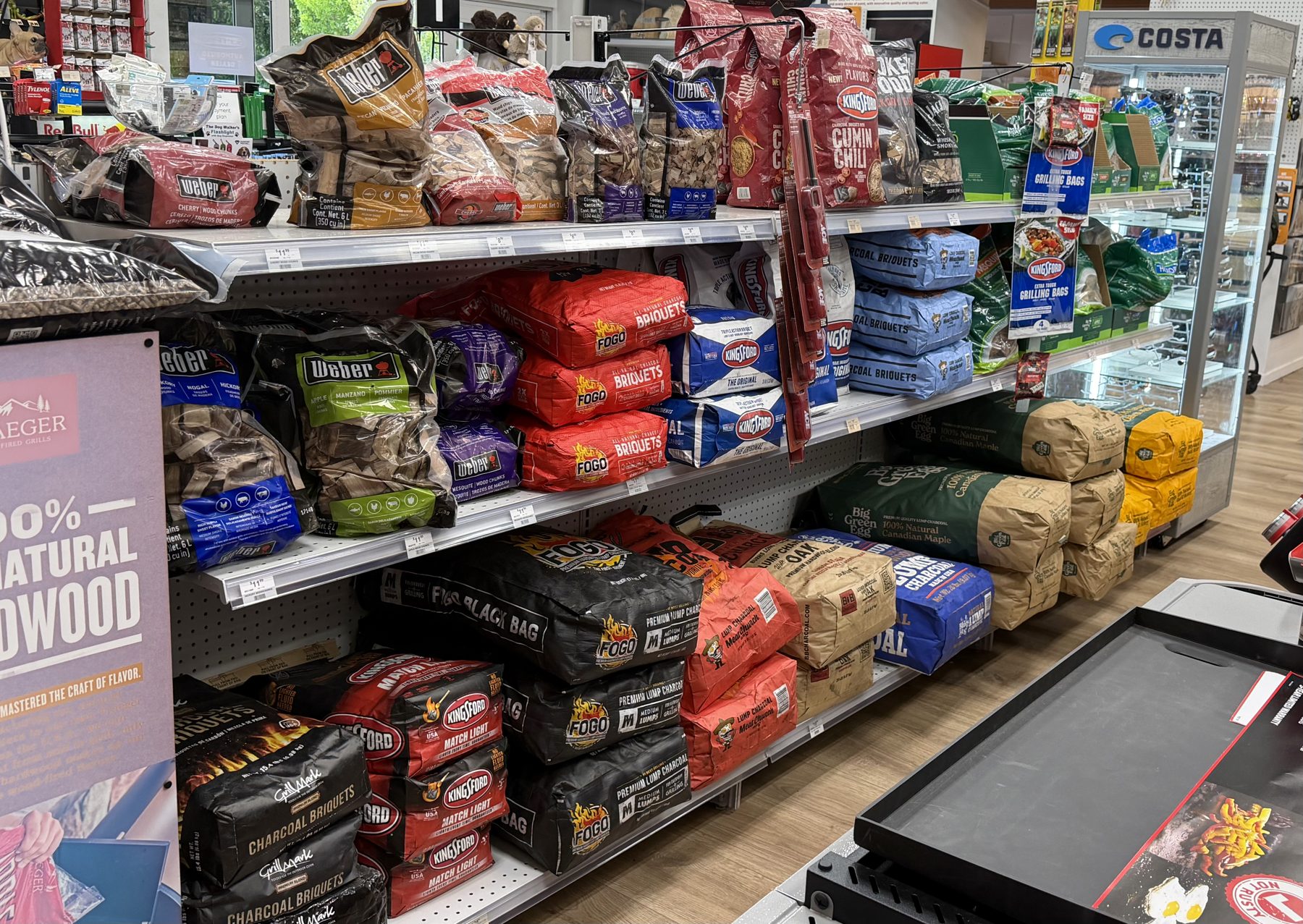 Shelves stocked with grilling fuel including Weber wood chunks, FOGO lump charcoal, Kingsford briquettes, and Big Green Egg natural charcoal at Hessler Paint & Hardware in Delray Beach, Florida.