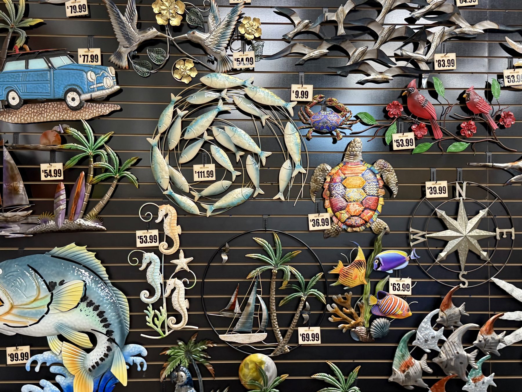 Display of decorative coastal metal wall art featuring sea turtles, schools of fish, seahorses, palm tree scenes, birds, and a metal compass rose hanging at Hessler Paint & Hardware in Delray Beach, Florida.