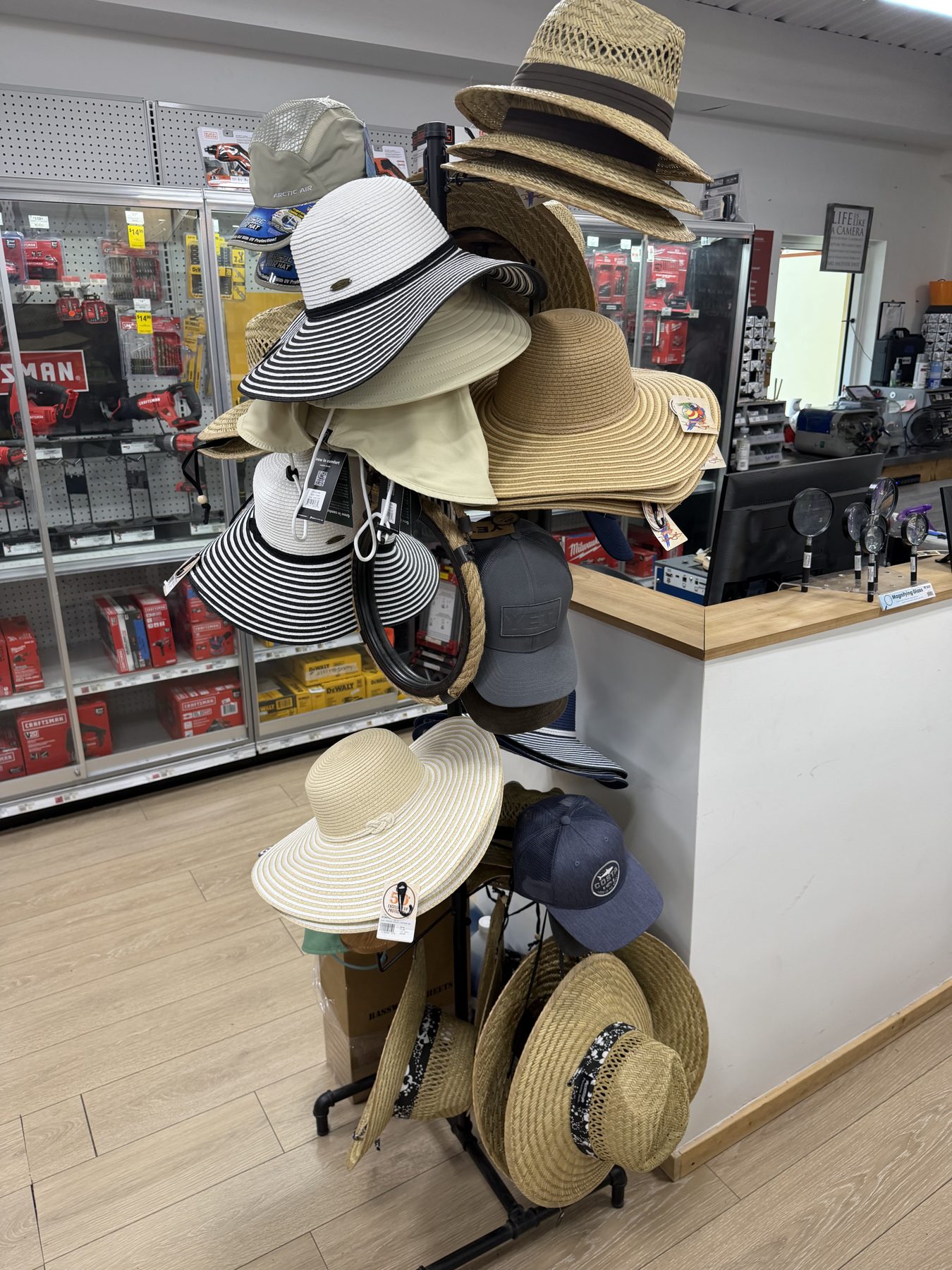 A freestanding display rack filled with wide-brim sun hats, straw garden hats, and baseball caps near the service counter at Hessler Paint & Hardware in Delray Beach, Florida.
