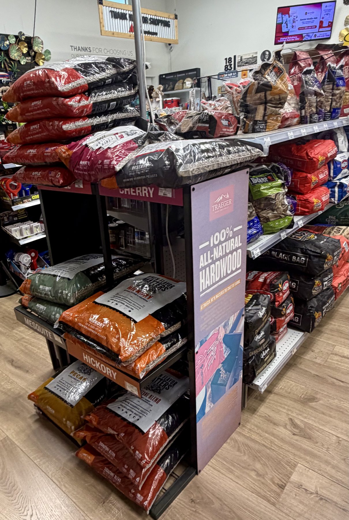 Traeger 100% all-natural hardwood grill pellets display featuring Apple, Cherry, Mesquite, Hickory, and Pecan flavors along with charcoal bags at Hessler Paint & Hardware in Delray Beach, Florida.