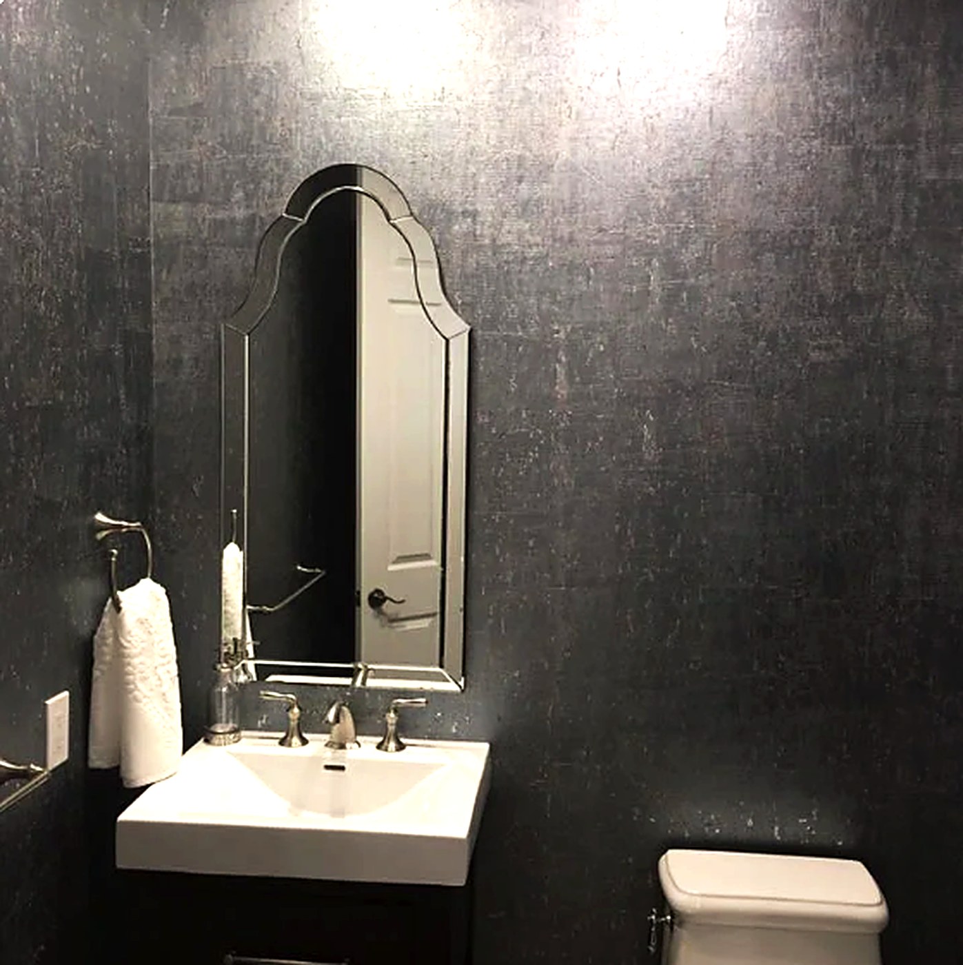 Powder room wrapped in dark charcoal metallic grasscloth wallcovering behind an arched mirror and wall-mount sink
