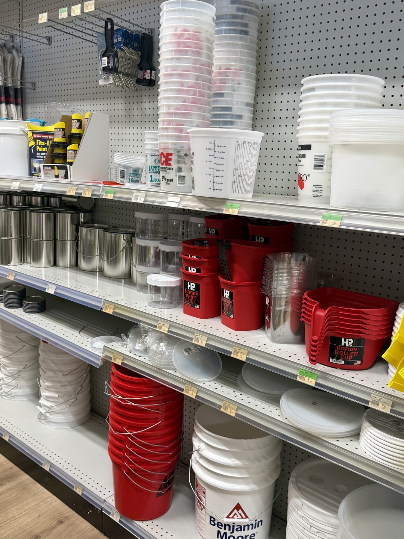 Aisle shelves fully stocked with Benjamin Moore 5-gallon pails, red HANDY Paint Pails and roller cups, metal paint cans, plastic bucket lids, and measuring containers at Hessler Paint & Hardware in Delray Beach, Florida.