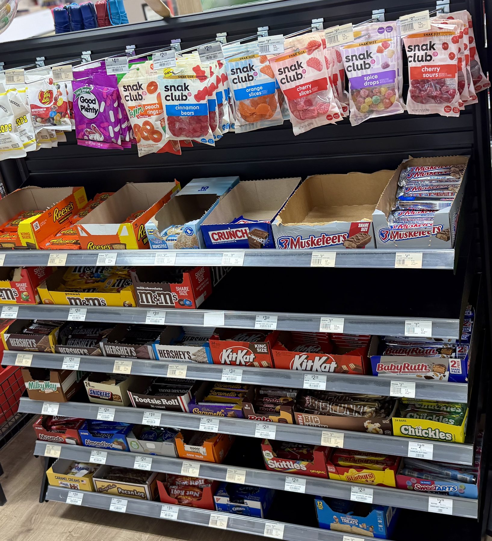 A shelf display filled with candy and snack items including Snak Club, Good & Plenty, Reese's, Crunch, 3 Musketeers, M&M's, KitKat, Hershey's, and Skittles at Hessler Paint & Hardware in Delray Beach, Florida.