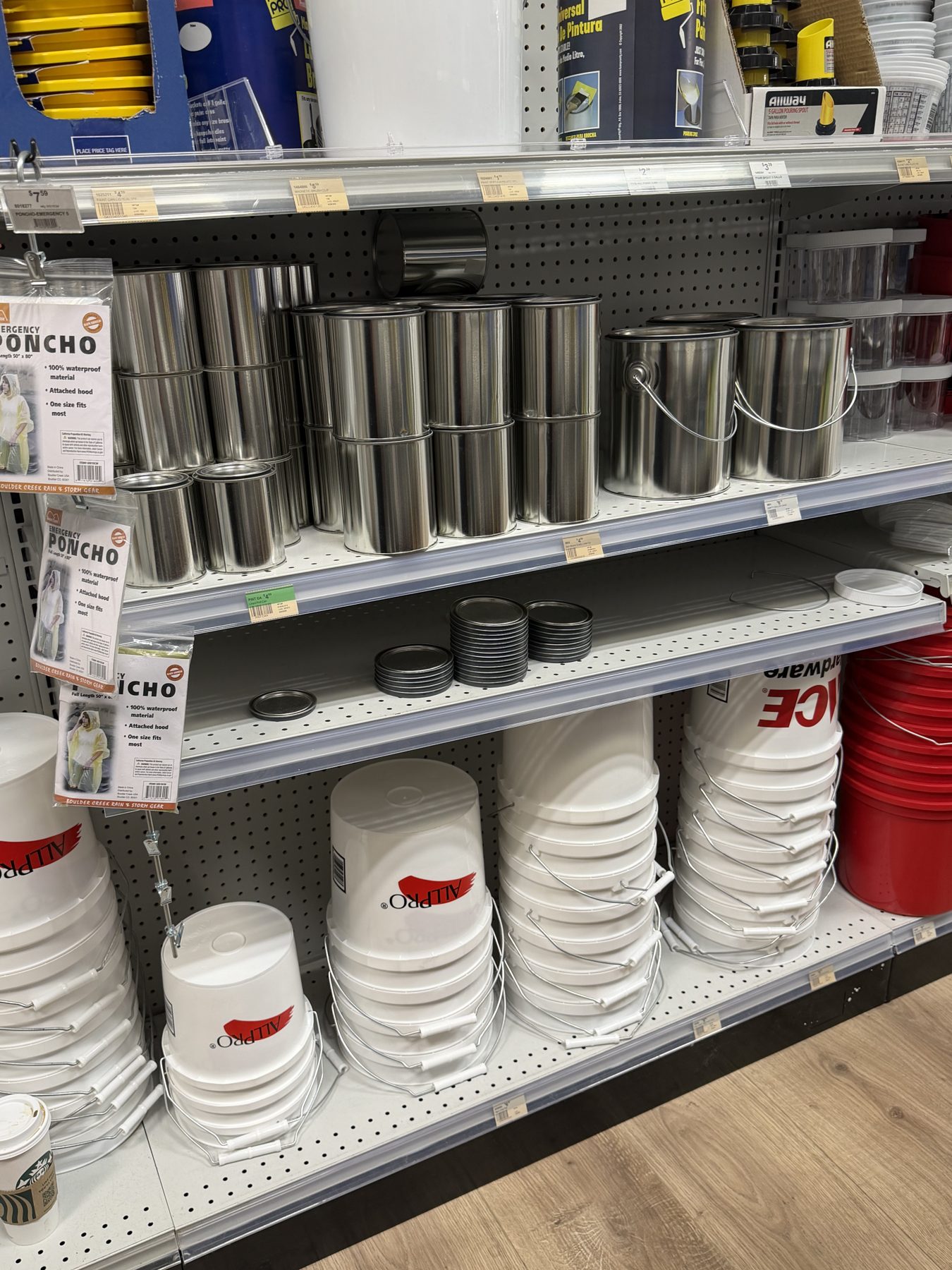 Shelves displaying empty metal paint cans, stacked ALLPRO white paint buckets, Ace Hardware buckets, paint lids, and hanging emergency ponchos at Hessler Paint & Hardware in Delray Beach, Florida.