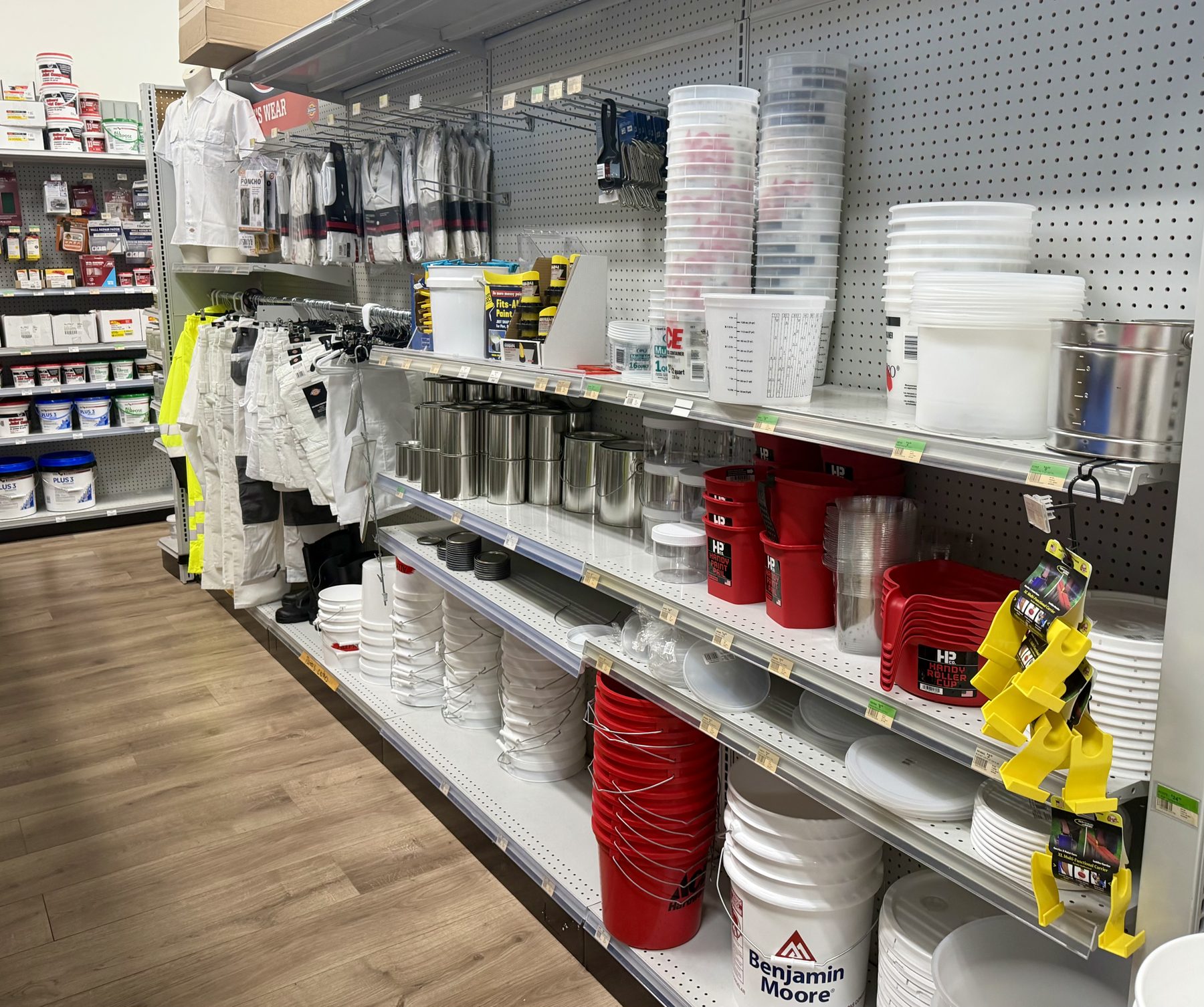 Shelves stocked with Benjamin Moore 5-gallon buckets, mixing containers, empty metal paint cans, Handy Paint Cup tools, and painter workwear at Hessler Paint & Hardware in Delray Beach, Florida.