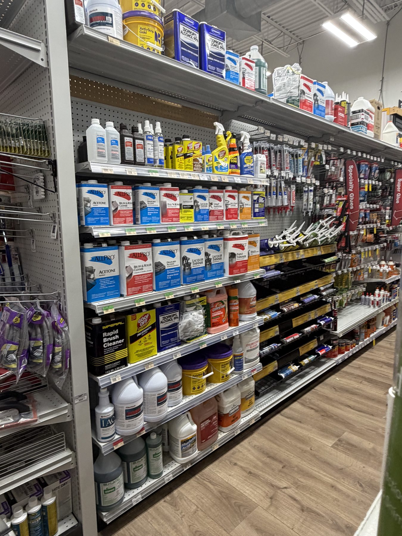 Aisle shelves fully stocked with Allpro acetone, lacquer thinner, paint thinners, Goof Off cleaner, and specialty glues at Hessler Paint & Hardware in Delray Beach, Florida.