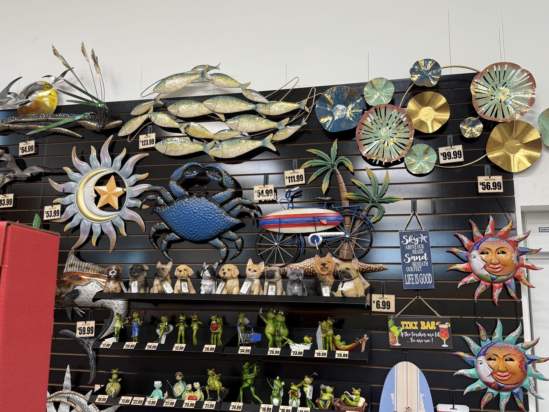 Vibrant coastal metal wall decor featuring a blue crab, school of fish, sunbursts, bicycle with surfboard, dog figurines, and frog statues on black slatwall shelves at Hessler Paint & Hardware in Delray Beach, Florida.