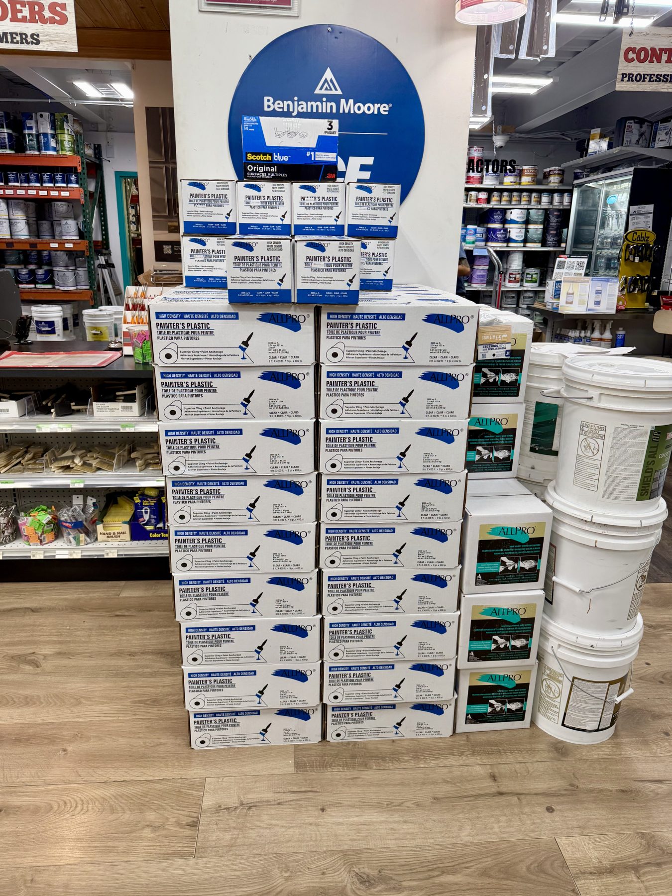 Stacked boxes of ALLPRO painter's plastic sheeting, ScotchBlue painter's tape, and 5-gallon paint buckets in front of a Benjamin Moore sign at Hessler Paint & Hardware in Delray Beach, Florida.
