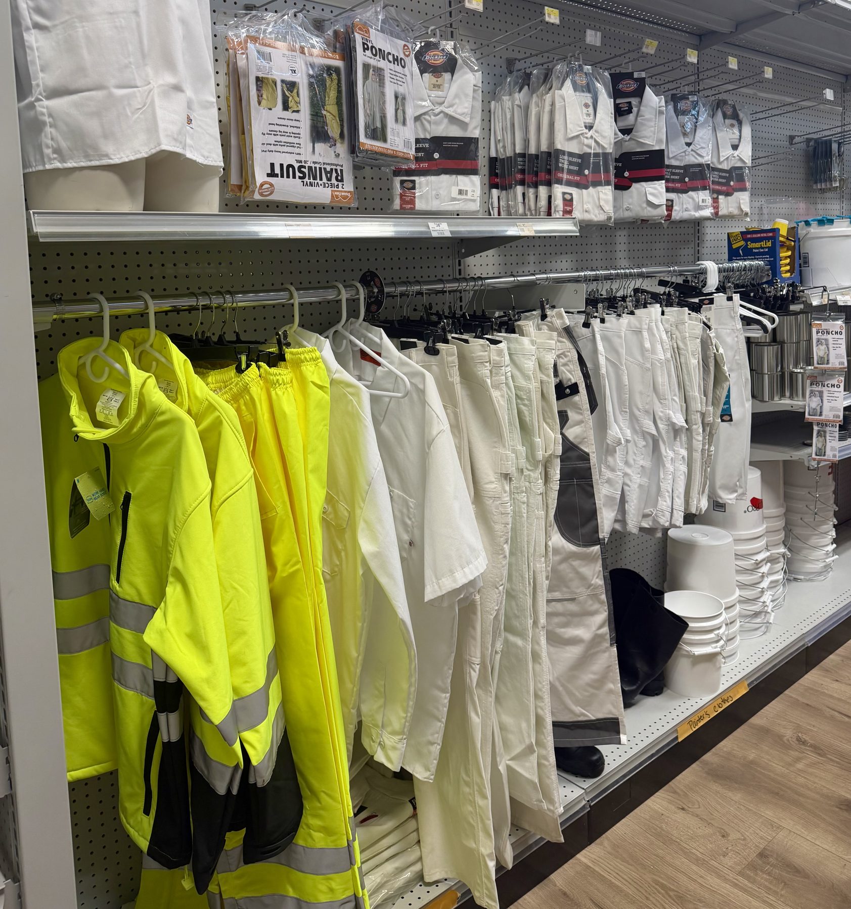 High-visibility safety jackets, Dickies painter pants, work shirts, rain suits, and paint buckets on retail shelves at Hessler Paint & Hardware in Delray Beach, Florida.