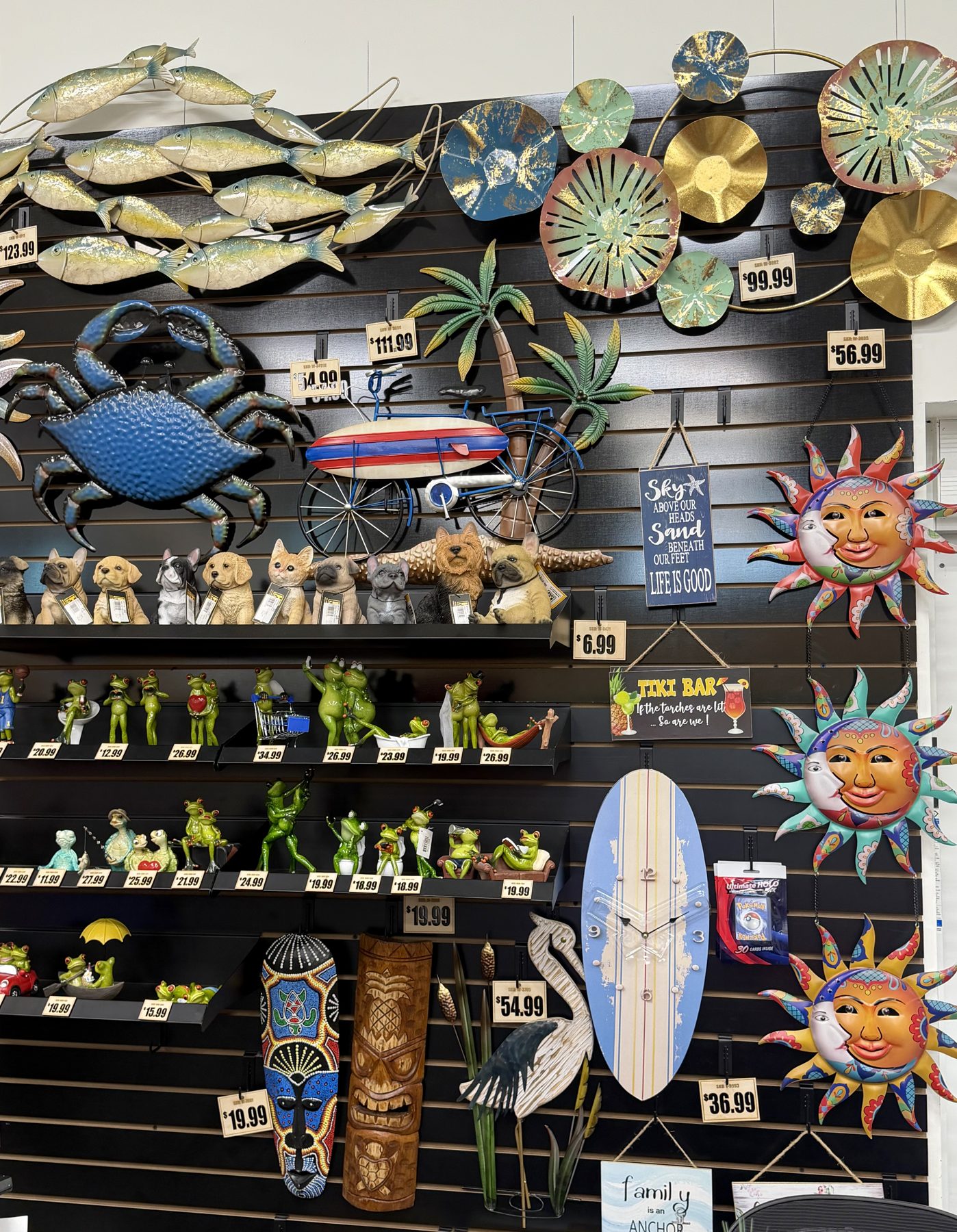 A vibrant display wall filled with tropical and coastal wall art, including metal fish, a blue crab, colorful sun face sculptures, tiki masks, frog figurines, and a surfboard clock at Hessler Paint & Hardware in Delray Beach, Florida.