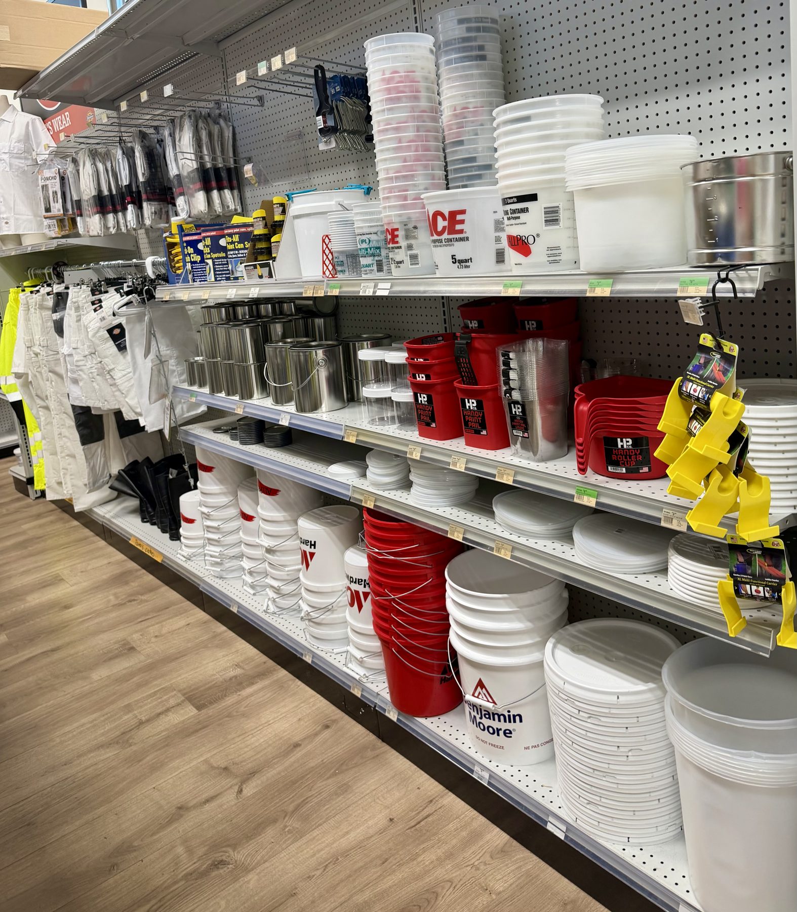 Display shelves stocked with Benjamin Moore and Ace Hardware 5-gallon paint buckets, plastic mixing containers, metal paint cans, Handy Paint Pails, and painter apparel at Hessler Paint & Hardware in Delray Beach, Florida.